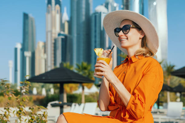 A woman in a bright outfit relaxes with a drink, with Dubai's skyline in the background.